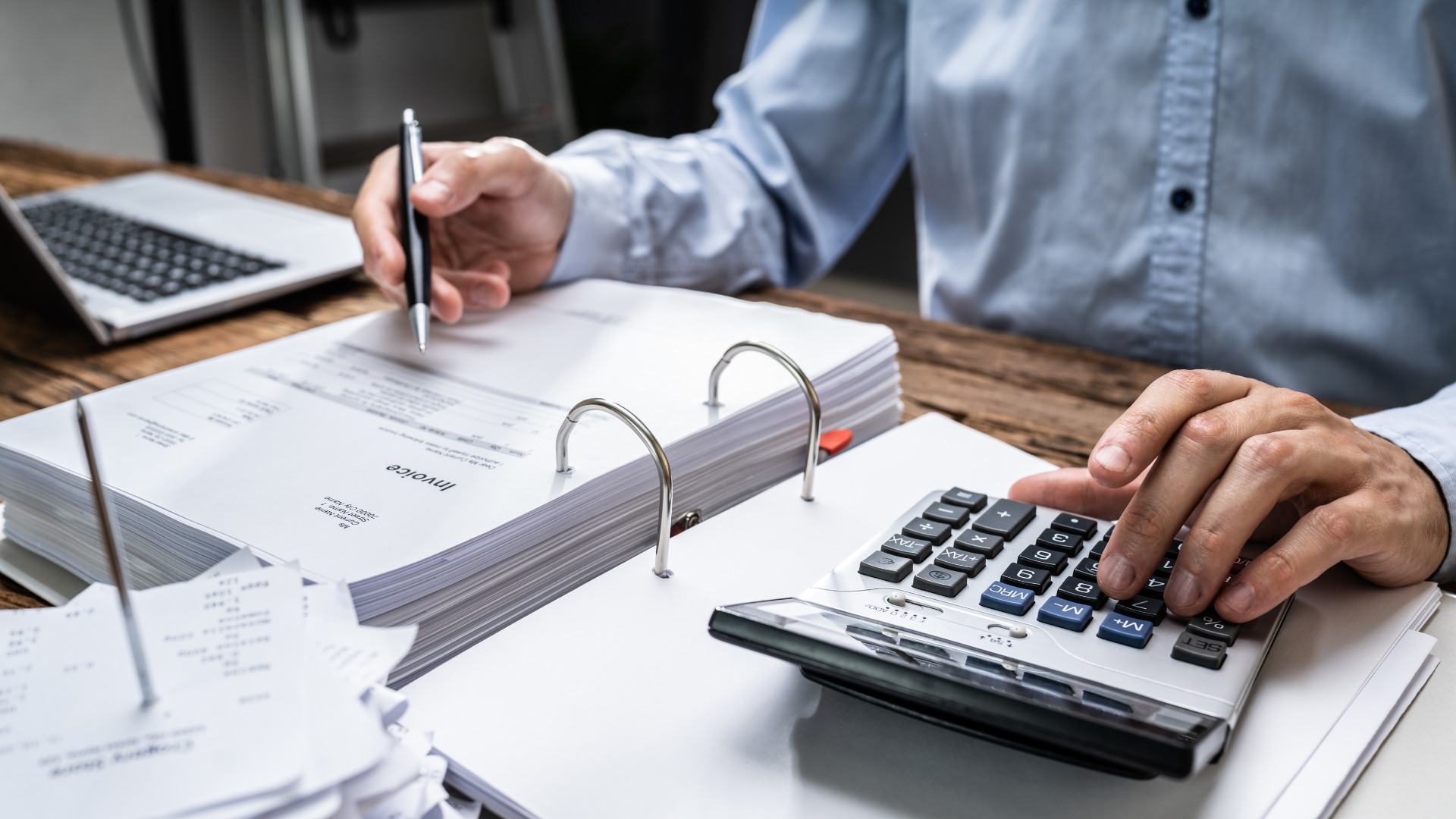 Personne travaillant sur des documents comptables avec une calculatrice et un classeur, sur un bureau en bois