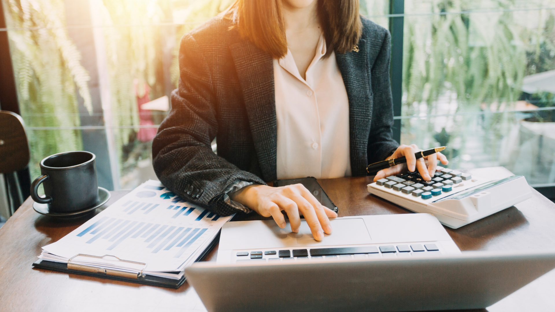 Femme utilisant une calculatrice et un ordinateur portable, travaillant sur des graphiques financiers à un bureau avec une tasse de café à proximité